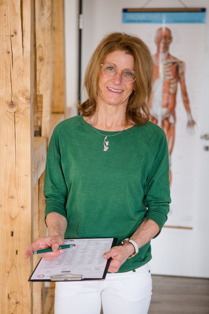 Tina Hirth mit einem Klemmbrett mit Daten. Im Hintergrund hängt ein Poster, das die Muskeln, Knochen und Organe eines Menschen zeigt. Tina Hirth bietet ein umfassendes Stoffwechselprogramm an.