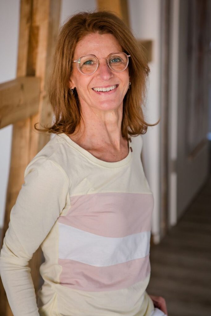 Tina Hirth steht im Praxisflur und lächelt in die Kamera. Sie macht mit ihren Kunden ganzheitliche Laboruntersuchungen.